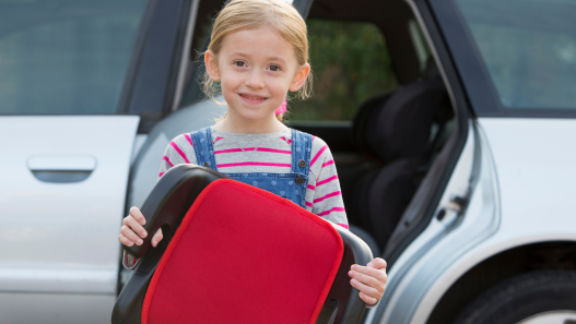 Child smiling in front of car holding a booster seat